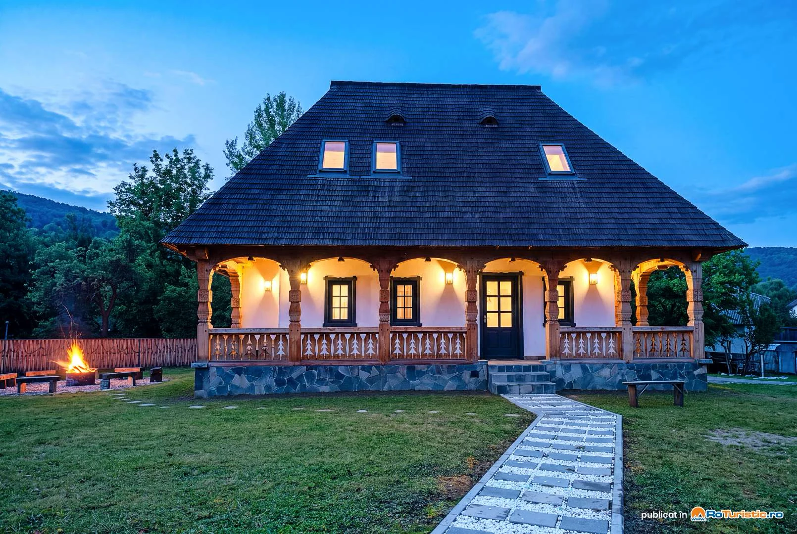 Chalet de Paul la lăsarea serii, cu prispa luminată și foc de tabără în curte